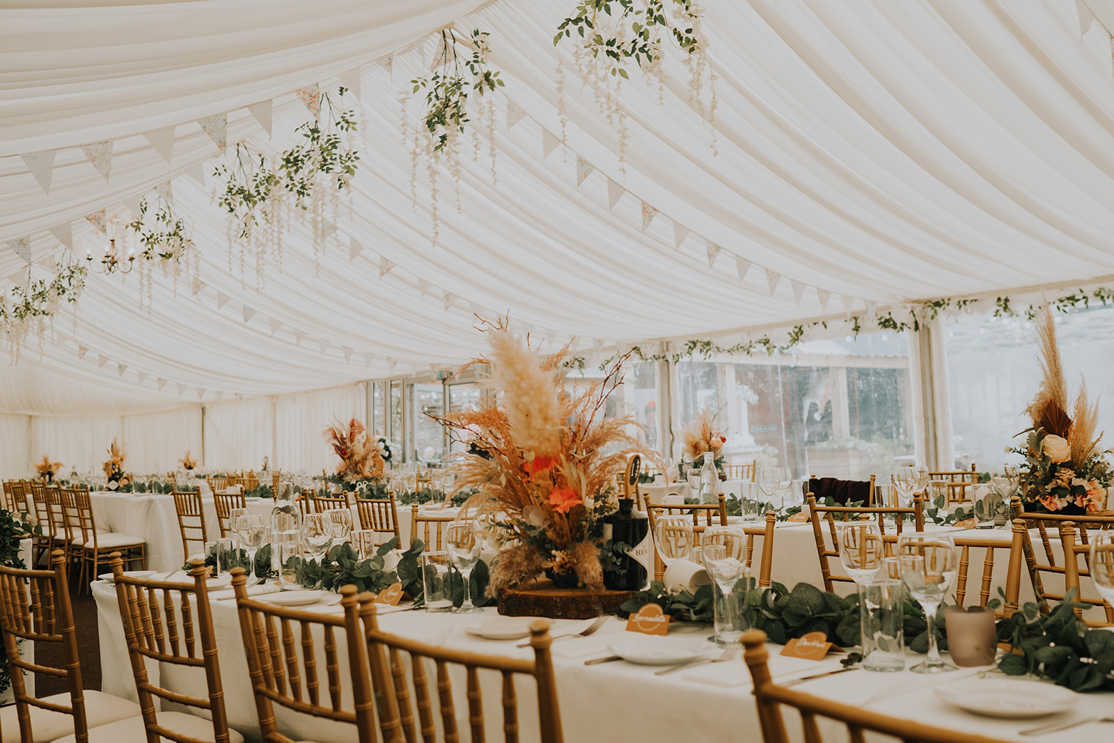 Draped marquee interior with long tables, gold chairs and dried floral centrepieces
