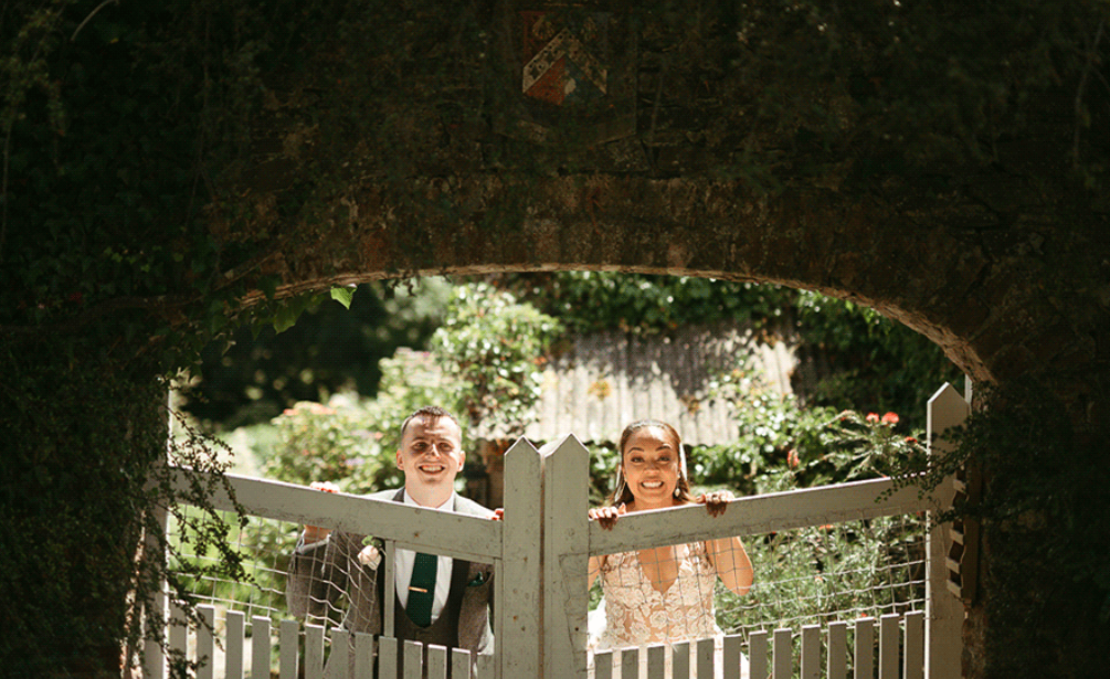 Bride and groom smiling at the white garden gate beneath the ivy-covered brick archway